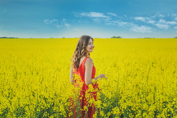 Fototapeta premium Young beautiful girl in a red dress close up in the middle of the yellow field with the radish flowers closeup
