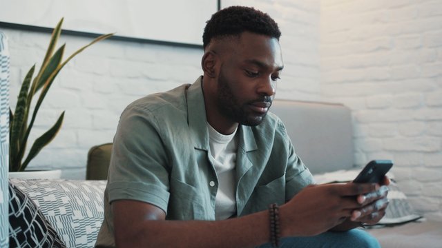 Handsome young african american man use phone lazy sit on sofa in home online relax work cell phone app authentic bills sunset guy close up mobile smartphone slow motion portrait handsome rude