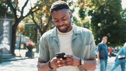 attractive african american young man walks down the street uses phone smile sunlight sunset cellphone fashion internet face outside technology black city mobile summer happy - Powered by Adobe