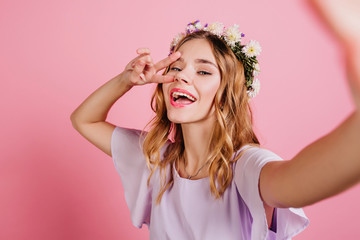 Inspired fair-haired girl in purple attire spending time in studio. Close-up portrait of lovable blonde lady in flower wreath making selfie and laughing.