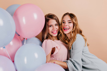 Laughing curly lady embracing her friend at party photoshoot. Portrait of girls with light-brown hair posing with colorful balloons on light background.