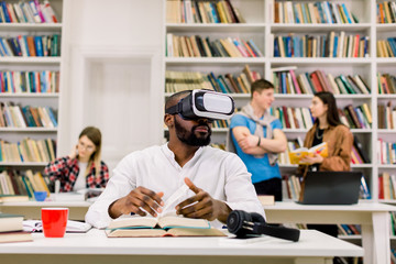 Black guy wearing vr goggles headset, reading book and using information from virtual reality, sitting in modern library. Group of students studying and talking each other on the background