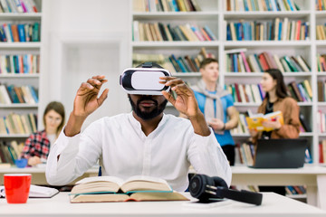 Male African student using VR goggles in public library and working with virtual textbook for...