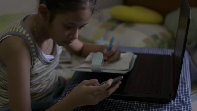 Asian cute girl in singlet shirt watching lesson online during doing homework on the bed. Female teenager studying from home with video call during COVID-19 outbreak situation.