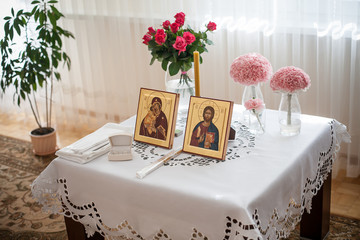 Orthodox wedding accessories. Saint portrait icons. Wedding rings, candles and flowers. White tablecloth ceremony preparation. Home interior important family holiday background.