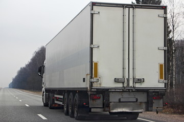 White semi truck close up with copy space blank on three-axle trailer rear door on empty highway road at spring day