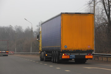 Semi truck with three-axle blue yellow trailer with copy space blank for text and logo on surfaces drive on suburban asphalted highway at spring day, international road transportation logistics