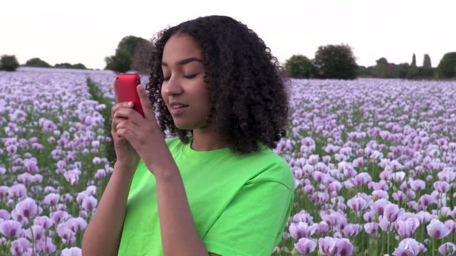 Beautiful happy mixed race African American girl teenager female young woman walking through field of pink poppy flowers taking photographs on her smart phone