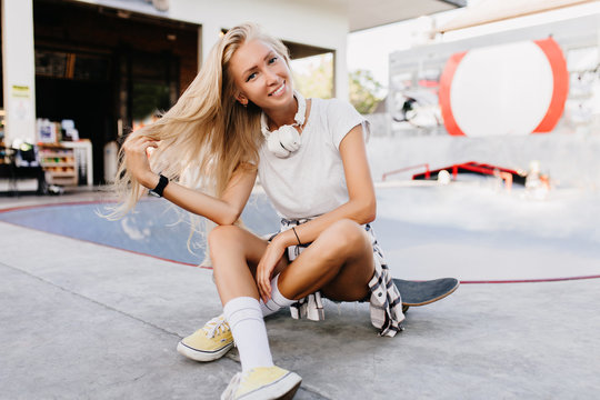 Pretty woman in yellow shoes sitting on longboard with legs crossed. Outdoor photo of stunning tanned female model having fun in skate park and playing with her hair.