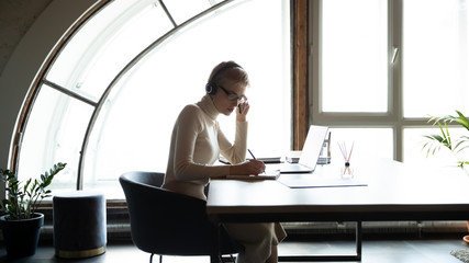 Side view concentrated 30s woman wearing headset with mic, studying remotely online alone at home, writing notes in organizer. Focused leader sitting at table, holding online meeting with coworkers.