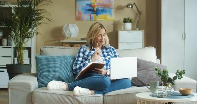Beautiful happy Caucasian woman working on laptop while sitting on sofa and making notes in planner at home. Pretty thoughtful female with computer writing down in notebook indoor. Freelance concept
