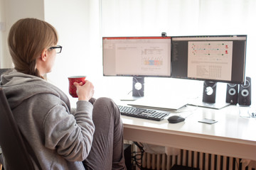 Unrecognizable Caucasian young woman scientist with glasses and coffee cup in casual home comfortable clothing working on computer with 2 monitors from home during covid-19 lock down