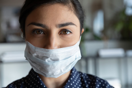 Close Up Head Shot Portrait Young Serious Cautious Indian Ethnic Woman Employee Worker Wearing Protective Medical Mask, Looking At Camera, Keeping Corporate Quarantine Rules, Healthcare Concept.