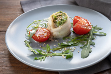 Fish fillet rolls from plaice filled with mashed celery and arugula, grilled tomatoes, rocket salad ande sesame seed on a gray plate, healthy appetizer suitable for Easter and spring time