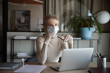 Distracted from job young thoughtful businesswoman in protective facial mask and gloves sitting at workplace, looking away, thinking of online company strategy development during quarantine time.