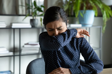 Millennial indian ethnic girl sitting in office, coughing in elbow, right illness behavior for not...