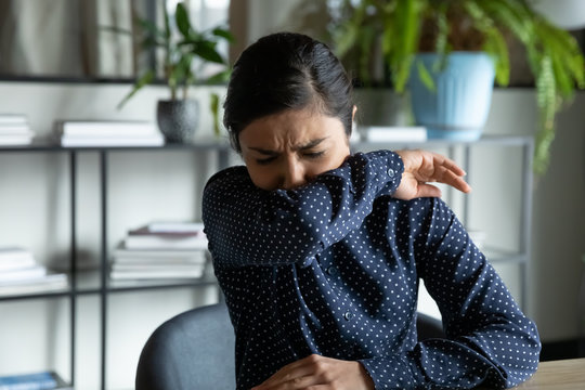 Millennial indian ethnic girl sitting in office, coughing in elbow, right illness behavior for not spreading virus infection. Unhealthy young woman feeling unwell at workplace, covid 19 symptoms.