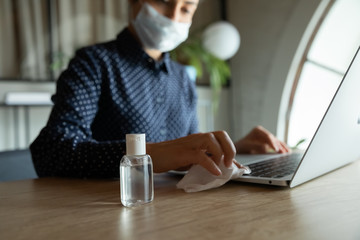 Focus on plastic bottle with antiseptic alcohol-based liquid standing on work desk with cautious woman in medical facemask sanitizing computer keyboard on background, stop spreading virus infection.