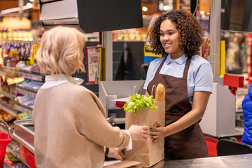 Pretty young cashier giving mature female paperbag with bread and groceries