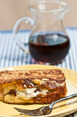 Top, front view, close distance of a maple, bacon, stuffed French toast, homemade, freshly baked, yellow, round plate, an artisan fork, glass pitcher of maple syrup, on blue, white stripped towel