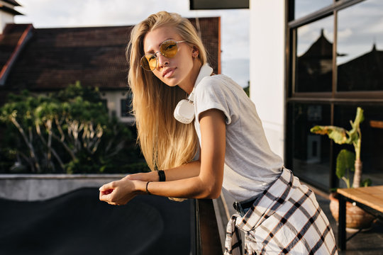 Good-looking blonde girl in trendy clothes posing with interested face expression in summer day. Photo of graceful caucasian woman in headphones and glasses standing outdoor.