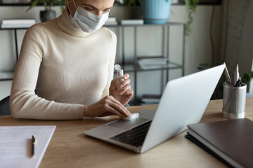 Cropped young 30s woman with short haircut wearing facial medical mask, wiping laptop keyboard and touchpad with antiseptic gel moistened napkin, working alone remotely from home, quarantine measures.