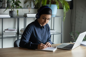 Smart millennial indian ethnicity woman sitting at desk with computer, wearing earphones, listening to educational seminar, attending online courses remotely from home, writing notes in copybook.