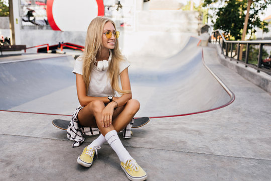 Gorgeous tanned skater girl posing outdoor. Photo of smiling female model in sport shoes sitting with legs crossed on skateboard.
