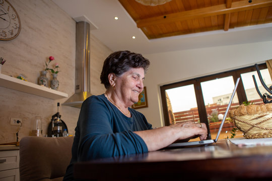 A Senior Woman With Black Short Hair Sits At A Dining Room Table And Works On A Laptop Computer. She Is Looking For Some New Cooking Recipes Online And She Is Thoughtful And Laughing.