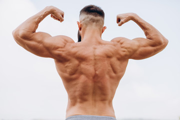 Handsome bearded bodybuilder man with perfect muscular body posing outdoors on the sky background. Strong man with naked torso standing, looking away. View from the back