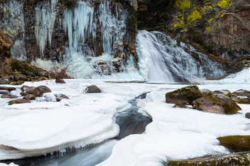 The Reinbach waterfall in Valle Aurina (Ahrntal), South Tyrol, Italy. White stalactites come down from the rocks, the water quickly pours into the small frozen lake. Granite and light green lichens.