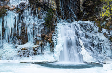 The Reinbach waterfall in Valle Aurina (Ahrntal), South Tyrol, Italy. White stalactites come down from the rocks, the water quickly pours into the small frozen lake. Granite and light green lichens.