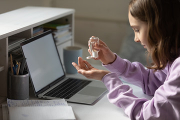 Young teenager schoolgirl cleaning hands with disinfecting antibacterial gel liquid, sitting at working table with computer during coronavirus quarantine time at home, starting doing homework.
