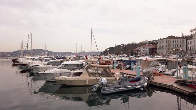 Cloudy foggy day. Private yachts and sailing boats with lowered sails standing on pier in bay. Reflections in water. People walk along promenade. Turkey, Istanbul, Tarabya, March 2020.
