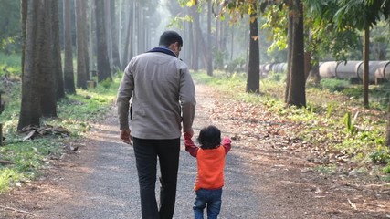 Backside of an Indian brunette father and his baby boy in winter garments walking in a forest path in winter afternoon in natural green background. Indian lifestyle and parenthood.