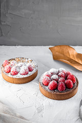 Appetizing tarts with dark chocolate, raspberries, crushed nuts and powdered sugar. Dry leaf near.  Natural light and color grade. White colored textured background. Copy space  