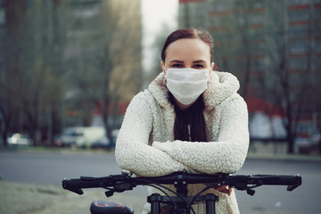 Young woman in medical mask stands with bicycle in city street. Adult female covered face with mask...