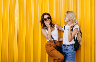 Stunning brown-haired girl in glasses laughing with blonde friend. Portrait of two young ladies spending summer vacation together and expressing positive emotions.