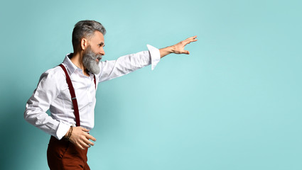 Elderly male in white shirt, brown pants and suspenders. He reaches for something by his hand, posing sideways on blue background