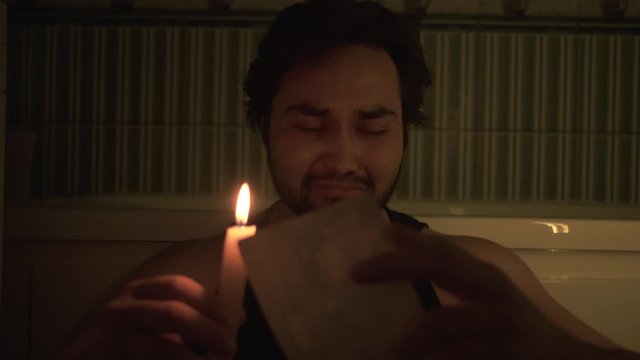 A desperate man reads a letter while sitting on the floor and holding a candle.