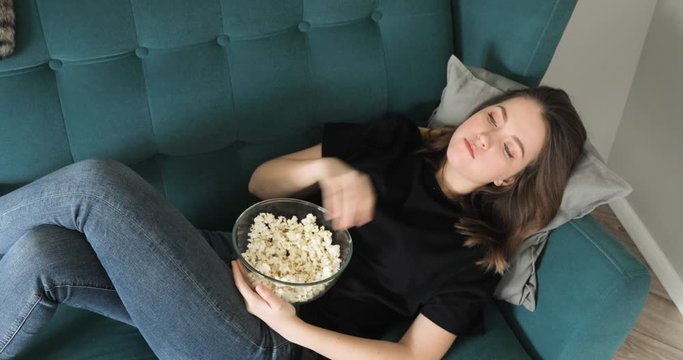 Top view of woman is eating popcorn lying on couch. Resting at home watching TV, movie, film, cinema. Movie food concept. Low calorie snack. Young girl in jeans and t-shirt.