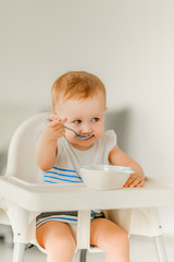 A little girl under the age of 1 year in a bright kitchen in a white highchair sits and eats from a white plate.