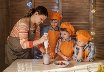 a mother and three sons prepare a milkshake with strawberries and a banana in the kitchen. boys dressed as cooks prepare dessert with their mother