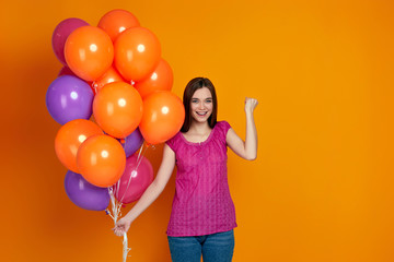 Fototapeta na wymiar Smiling delighted girl in pink t-shirt with bright colorful air balloons raises clenched fists isolated over orange background. Beautiful happy young woman on a birthday party. space for text