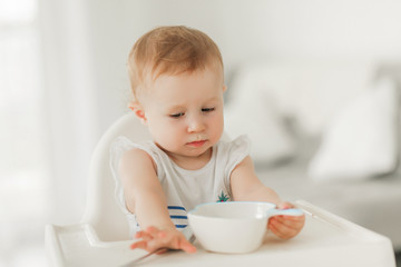 A little girl under the age of 1 year in a bright kitchen in a white highchair sits and eats from a white plate.