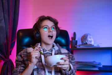 Excited young woman watching video on a computer and eating snack. Neon light at the background.