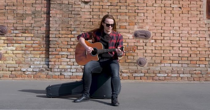 Young male with long hair and tattoo play on acoustic guitar outdoors on the street	