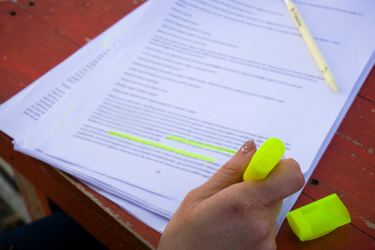 Female hand holding highlighter pen, highlighting / marking important texts in study sheets on table background.