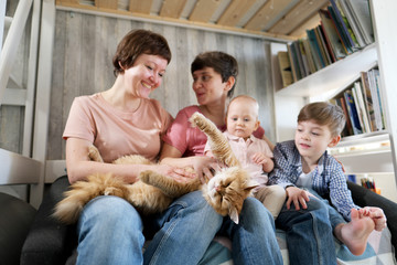 A young lesbian same-sex family with two children, a son and a daughter spend time at home. They sit on the couch and read a book. The pet cat is lying next to it.