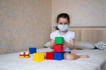 a little girl in a protective mask plays with educational toys, sitting on the bed in self-isolation, look at camera